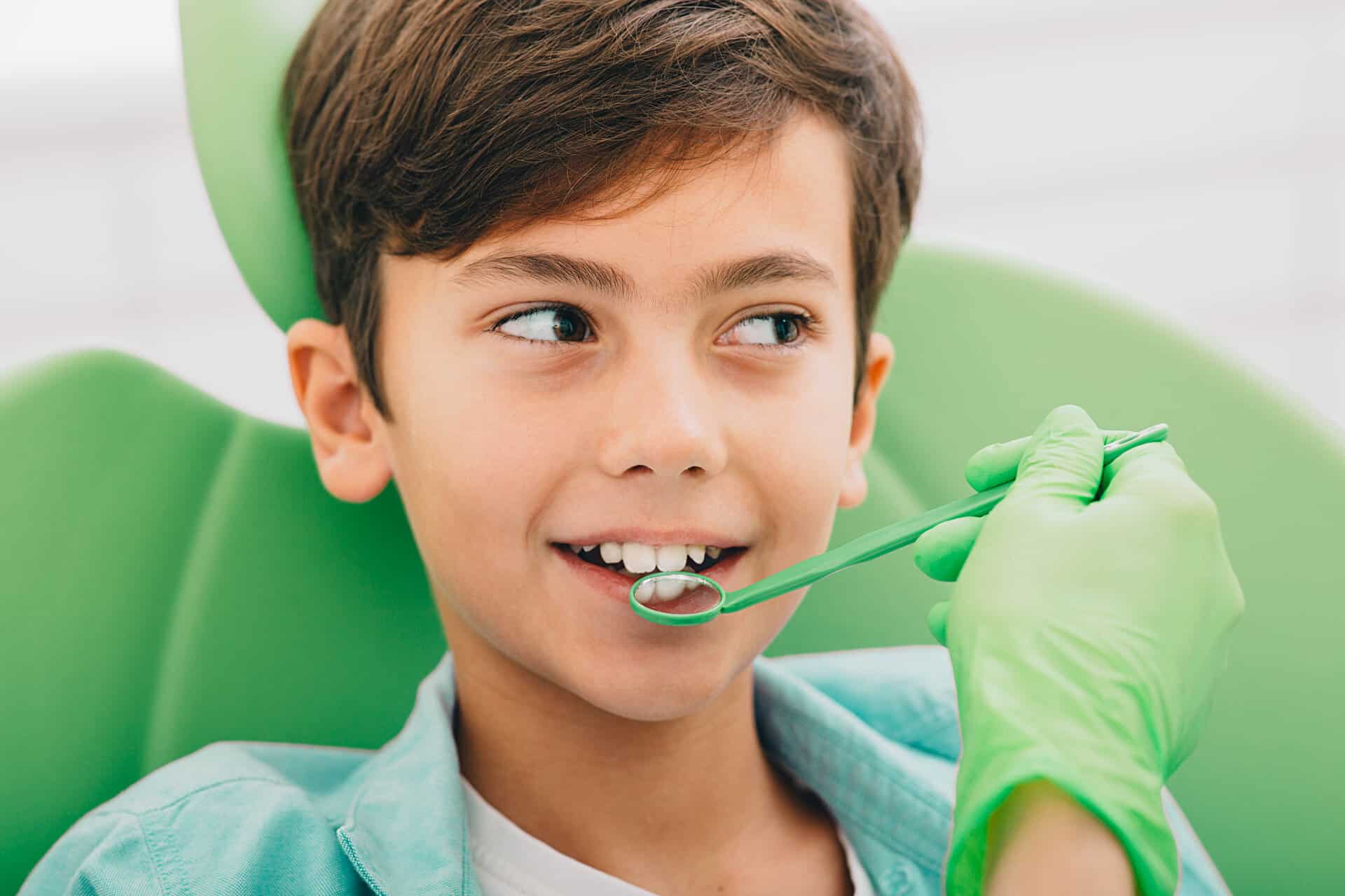 A child in a dental chair smiles while a person wearing green gloves holds a dental tool near the child’s mouth.