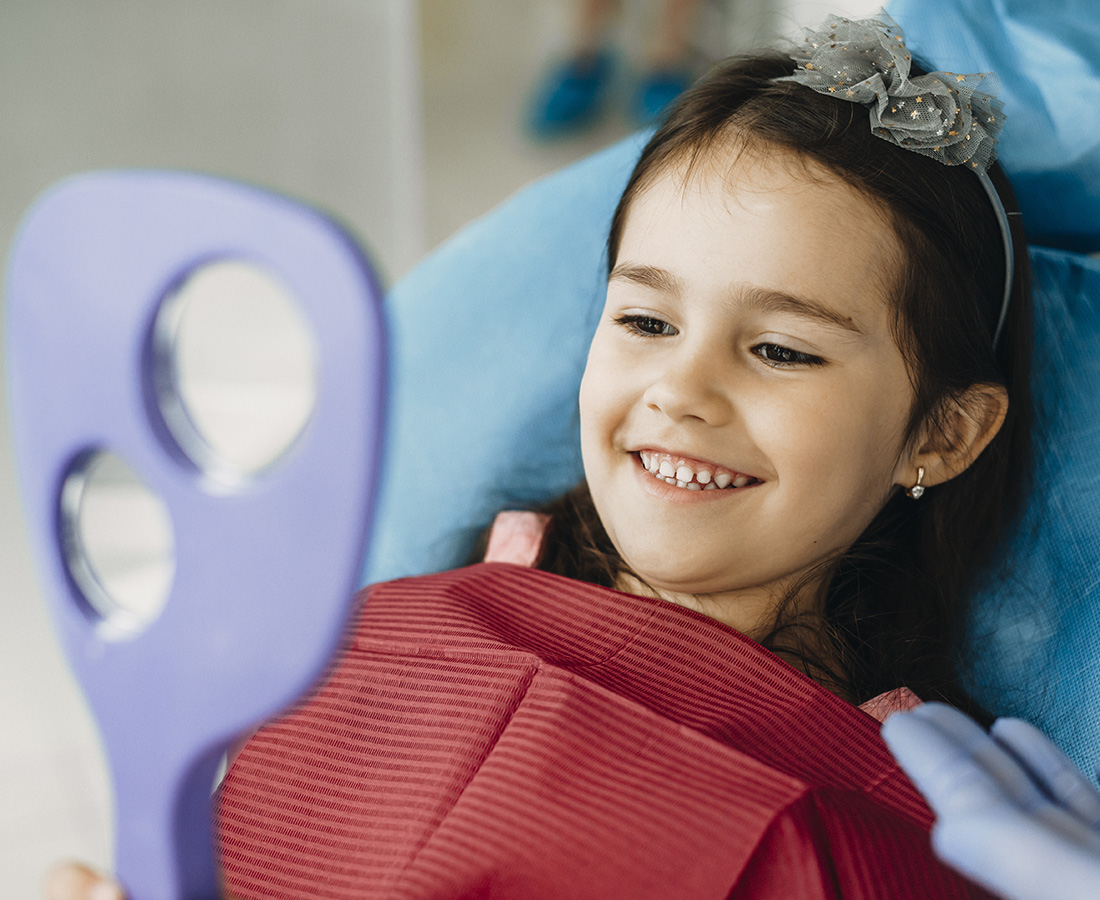 A child with a dental bib smiles while holding a hand mirror, reflecting a joyful expression during a dental check-up.