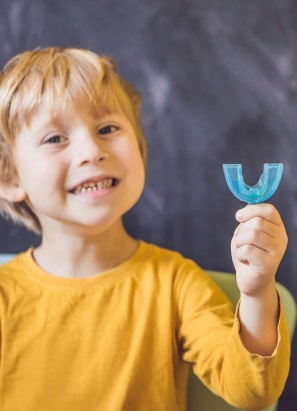 A child wearing a yellow shirt smiles while holding a blue dental retainer in their hand. A chalkboard background is visible behind the child.