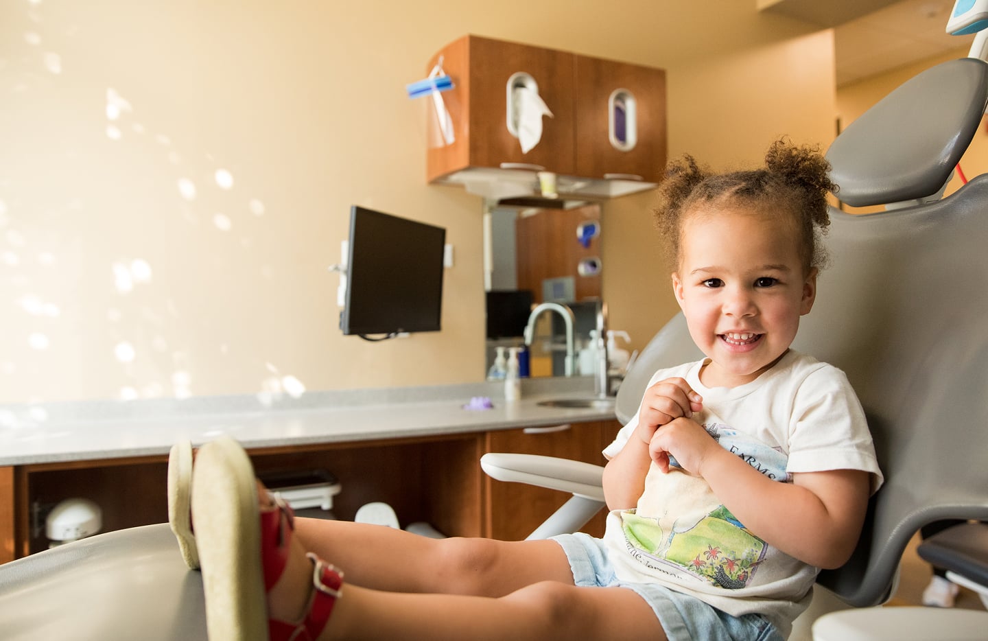 A child sits smiling in a dental office, seated on a dental chair, with medical equipment and wooden cabinets in the background.