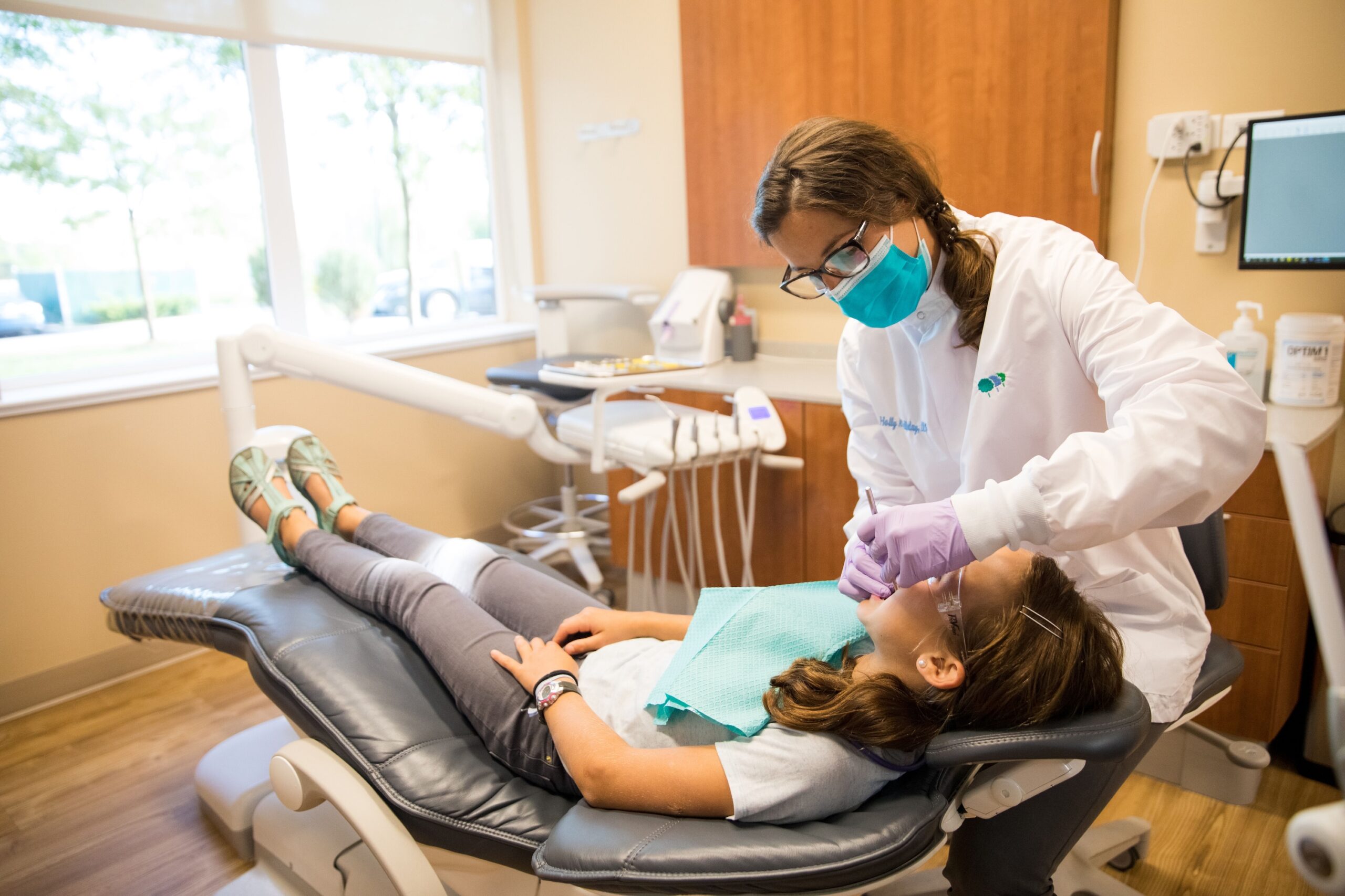 A person in a dental office is examining another person lying on a chair, with medical equipment visible around them.