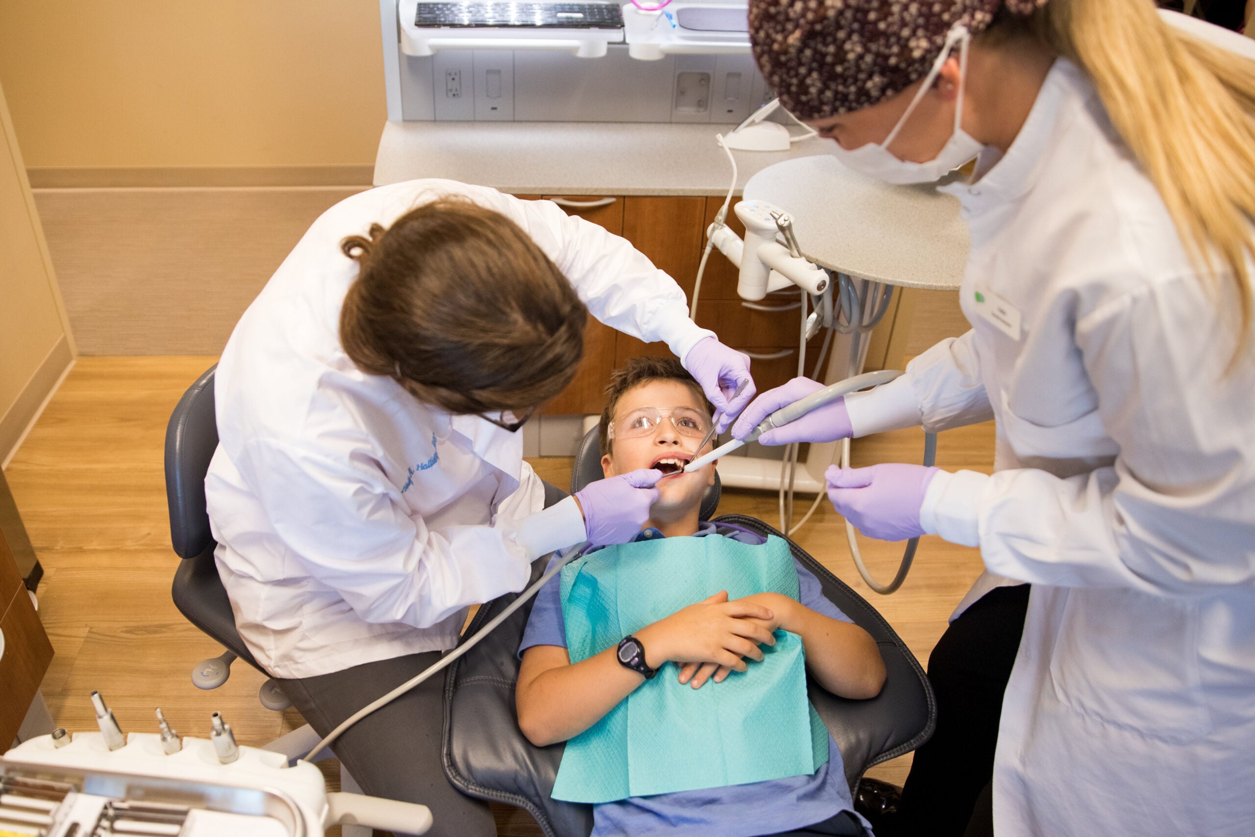 A child is receiving dental treatment from two persons wearing masks and gloves in a clinic. Dental equipment is visible around them.