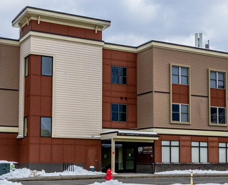A modern, three-story building with a mix of beige and reddish-brown panels, surrounded by snow, and featuring multiple windows and an entrance.