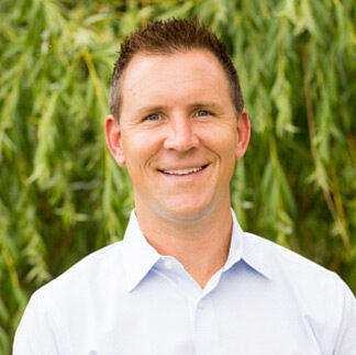 A person in a white shirt stands smiling in front of green foliage, suggesting an outdoor setting. No landmarks or historical buildings visible.