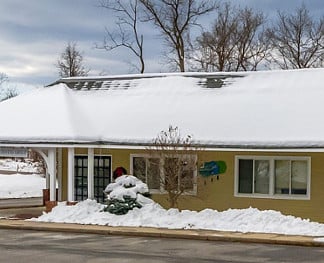 Snow-covered building with light yellow siding, surrounded by bare trees and snow piles. Overcast sky above, creating a wintery scene.