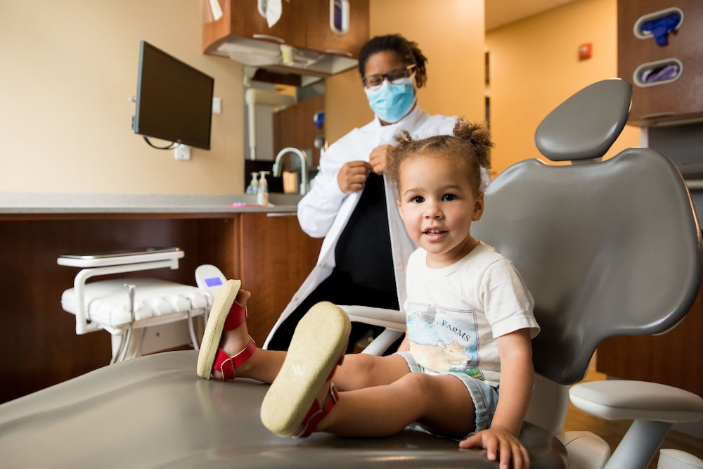 A child sits on a dental chair, smiling, while a person in a white coat prepares equipment in a dental office.