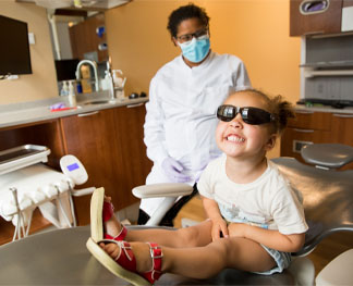 A person in a dental office and a child wearing sunglasses, smiling while sitting on a dental chair. Medical equipment visible in background.